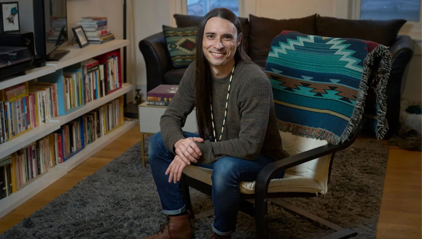 Environmental portrait of Joseph Pierce sitting in a room with a bookshelf in the background.